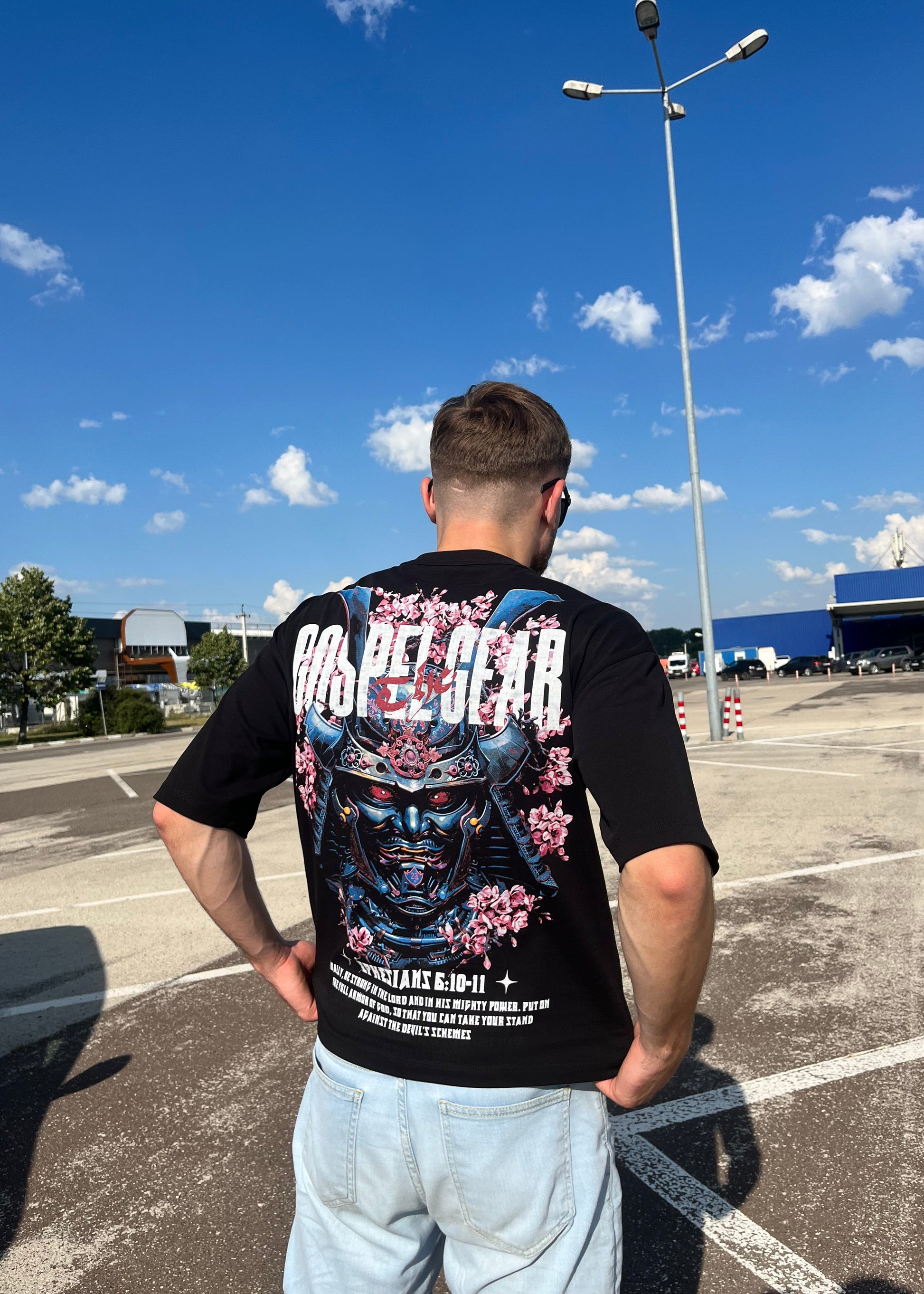 Person wearing a black t-shirt with colorful graphics and text, standing on a street with a clear blue sky.